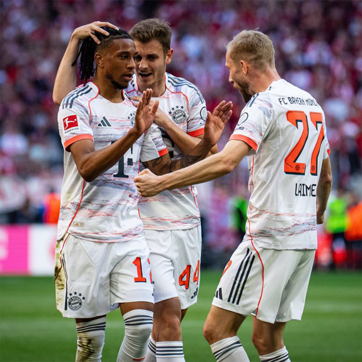 Three soccer players from Bayern Munich celebrate a goal on the field. One player, wearing jersey number 14, is being congratulated by teammates as they express excitement and camaraderie. The background shows a crowd, emphasizing the atmosphere of a match.