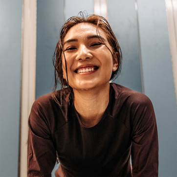 A smiling young woman with damp hair leans forward, conveying a sense of joy and satisfaction. She wears a dark athletic shirt and stands against a light blue background. Her expression suggests confidence and contentment, possibly after a workout or physical activity.