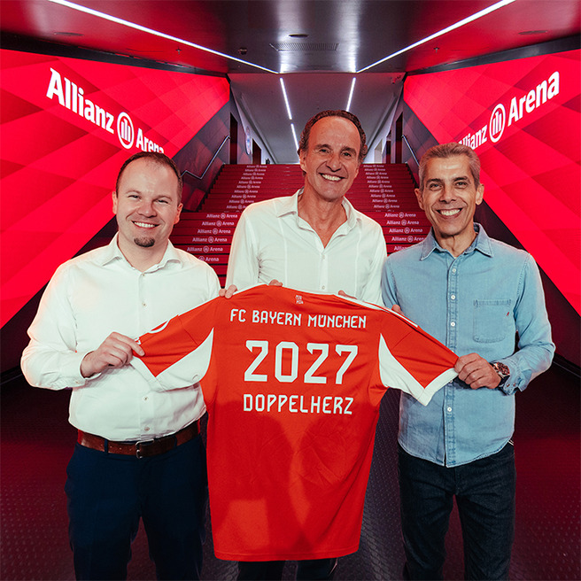 Three men stand together in a corridor of Allianz Arena, holding up a red FC Bayern München jersey with "2027" and "DOPPELHERZ" printed on it. The background features red lighting and stair markings, emphasizing the venue's branded atmosphere.
