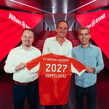 Three men stand together in a corridor of Allianz Arena, holding up a red FC Bayern München jersey with "2027" and "DOPPELHERZ" printed on it. The background features red lighting and stair markings, emphasizing the venue's branded atmosphere.