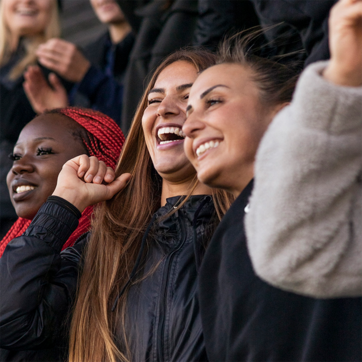 Three women are joyfully cheering and smiling in a lively setting, with one woman gleefully raising her hand. They appear to be celebrating, conveying a sense of happiness and excitement. Their expressions highlight a moment of shared enthusiasm.